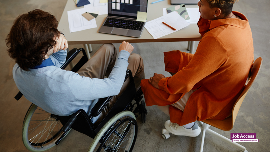 Person in a wheelchair with another person in an orange top at a desk working on a computer.
