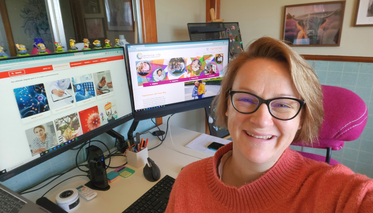 Chelsea is sitting in a chair, smiling at the camera. She is wearing glasses and an orange jumper. Two computer monitors and a work desk are behind her.