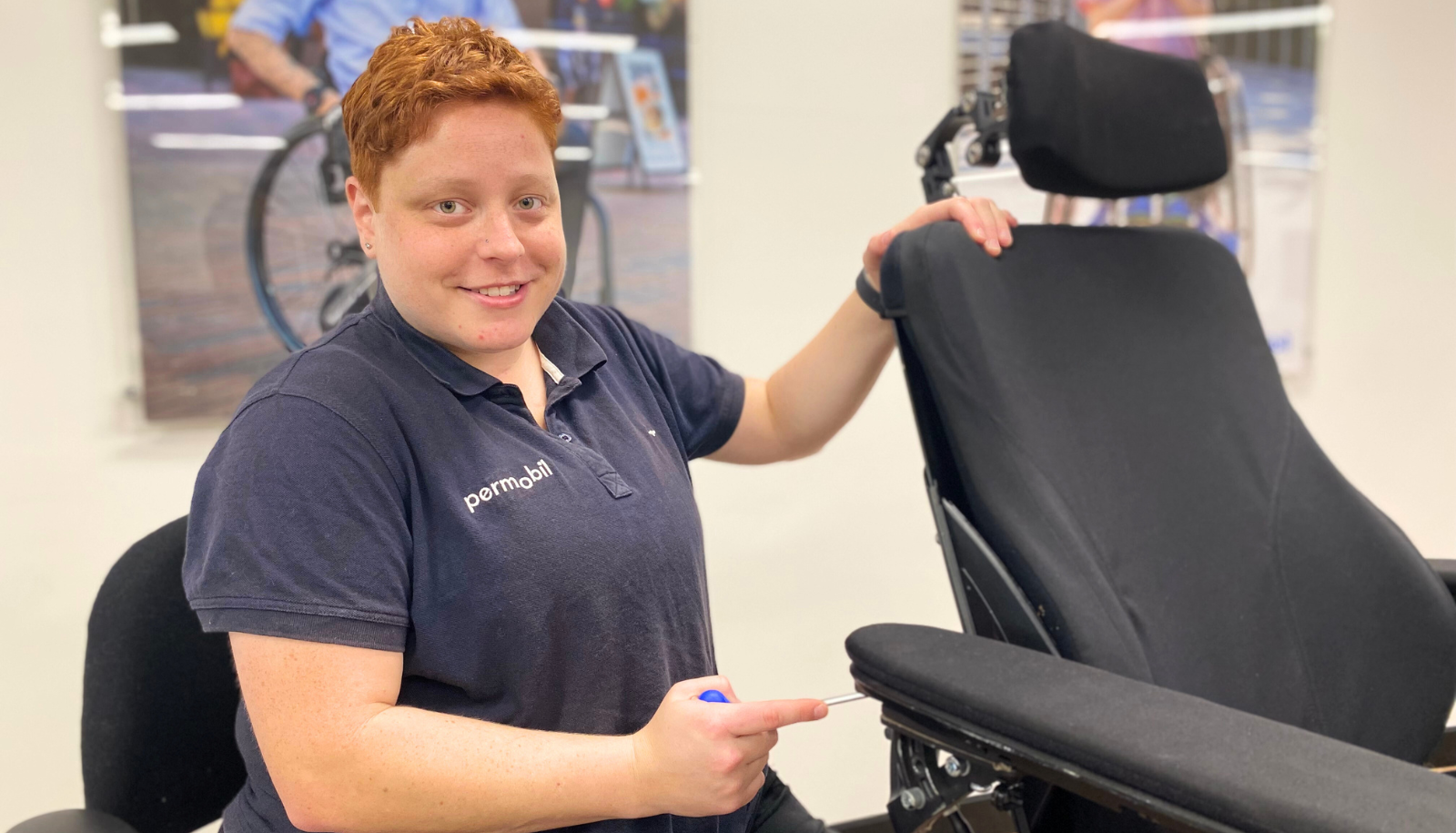 Monique sits in her ‘magic chair’. She wears a navy-blue polo shirt and smiles at the camera. Monique adjusts the armrest width, made easier on her body by elevating the height of the ‘magic chair’ seat so her torso and shoulders aren’t strained.
