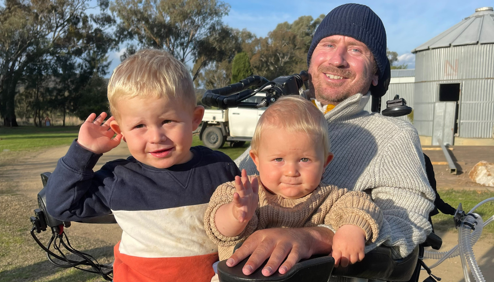 Nathan Stapleton is in a wheelchair outdoors at a farm with his two sons sitting on his lap