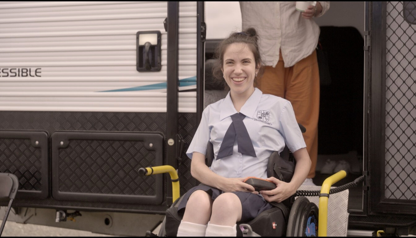 Hannah Diviney is sitting in her accessible wheelchair on the sets of ‘Audrey’. She is wearing a light-blue shirt with a criss-cross bow tie and smiling at the camera. There is wheelchair-accessible caravan behind her.