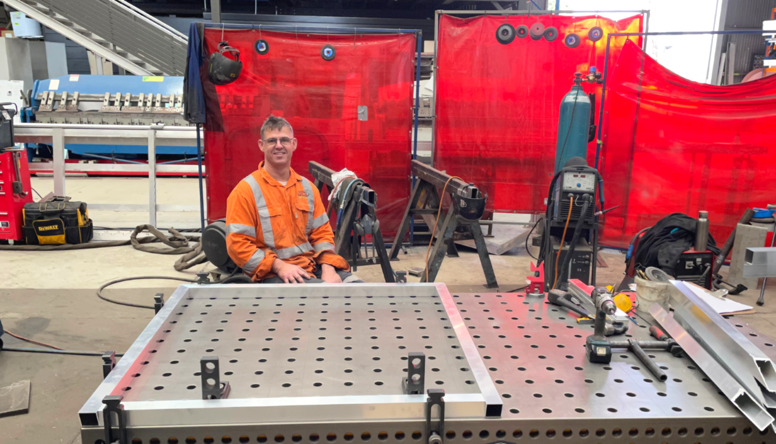 Image of Clay is sitting on a perching stool in the factory and smiling at the camera. He is surrounded by factory equipment and seated behind a three-dimensional (3D) welding table.