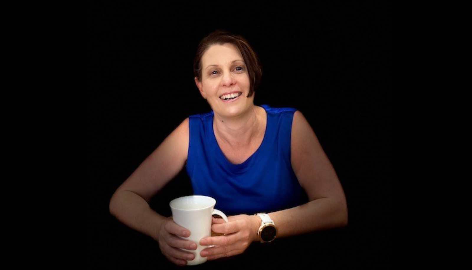Sarnya is sitting while smiling to camera, wearing a blue top and holding a white mug.